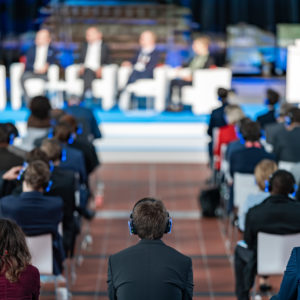 Foto eines Event-Teilnehmers, der Kopfhörer trägt. Im Hintergrund findet auf der Bühne eine Talkrunde statt